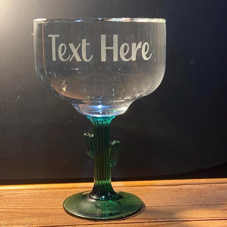 May include: A clear glass margarita glass with a green cactus stem and base. The text "Text Here" is etched on the glass. The glass is designed to hold a generous serving of a margarita or other cocktail.