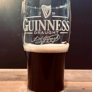 May include: A pint glass of Guinness stout, with a thick, creamy head. The glass is etched with the Guinness logo and the words "Guinness Draught". The dark beer contrasts with the white foam. The background is dark.