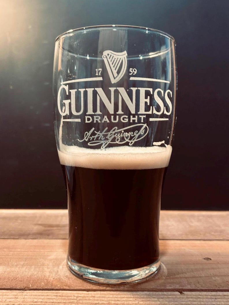 May include: A pint glass of Guinness stout, with a thick, creamy head. The glass is etched with the Guinness logo and the words "Guinness Draught". The dark beer contrasts with the white foam. The background is dark.