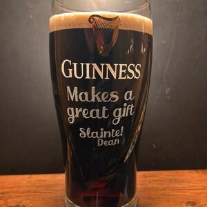 May include: A pint glass filled with dark stout, featuring a creamy head. The glass is etched with the Guinness logo and the words "Guinness Makes a great gift. Sl&aacute;inte! Dean."