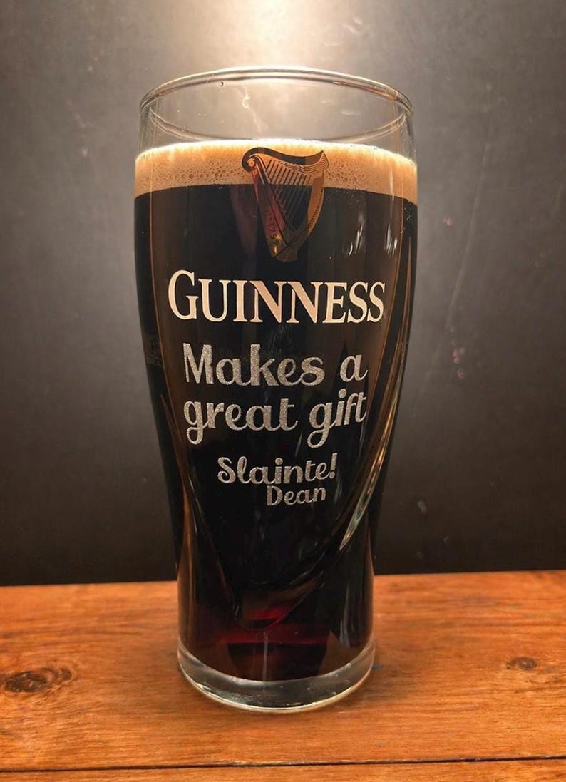May include: A pint glass filled with dark stout, featuring a creamy head. The glass is etched with the Guinness logo and the words "Guinness Makes a great gift. Sl&aacute;inte! Dean."
