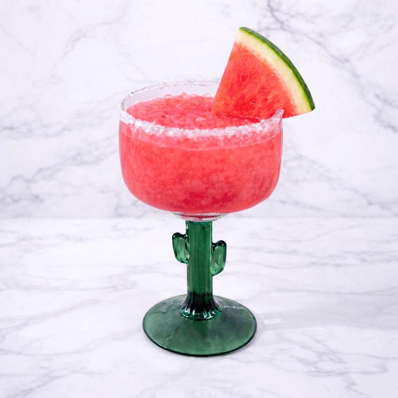 Pu&ograve; includere: Un rinfrescante margarita all'anguria in un bicchiere verde a forma di cactus. Il bordo &egrave; ricoperto di sale e una fetta di anguria adorna la bevanda. Su sfondo di marmo bianco.