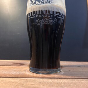 May include: A pint glass of dark stout beer with a thick, creamy head. The Guinness logo is etched on the glass, with the word "Draught" and Arthur Guinness' signature. The glass is on a wooden surface, with a dark background.