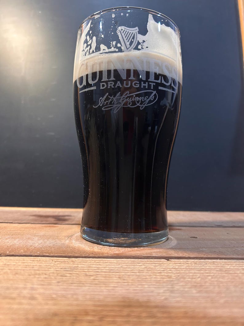 May include: A pint glass of dark stout beer with a thick, creamy head. The Guinness logo is etched on the glass, with the word "Draught" and Arthur Guinness' signature. The glass is on a wooden surface, with a dark background.