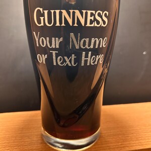 May include: A pint glass of dark stout beer with a thick, creamy head. The glass is branded with the Guinness logo and the text "Your Name or Text Here" in silver. The glass is resting on a wooden surface.