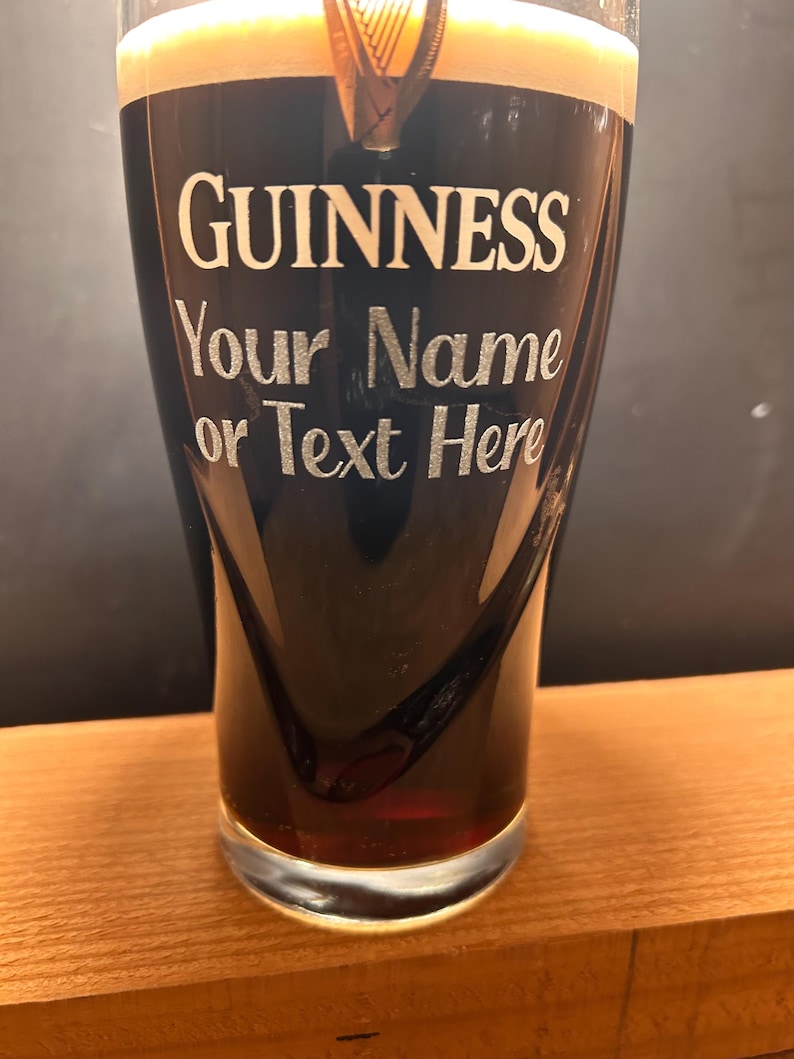May include: A pint glass of dark stout beer with a thick, creamy head. The glass is branded with the Guinness logo and the text "Your Name or Text Here" in silver. The glass is resting on a wooden surface.