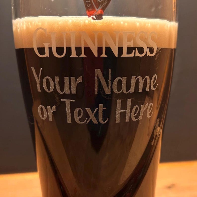 May include: A pint glass of dark stout beer with a creamy head. The glass is etched with the word "GUINNESS" and the text "Your Name or Text Here". The glass is dark with a curved design.