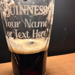 May include: A pint glass with the Guinness logo and the text "Your Name or Text Here" etched on the front. The glass is filled with a dark stout, topped with a creamy, off-white foam.