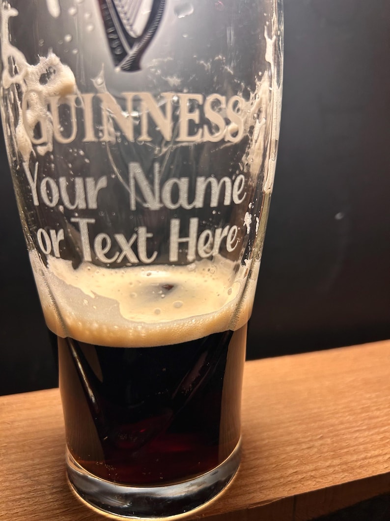 May include: A pint glass with the Guinness logo and the text "Your Name or Text Here" etched on the front. The glass is filled with a dark stout, topped with a creamy, off-white foam.
