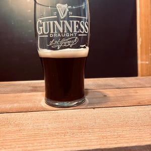 May include: A pint glass of dark stout beer with a creamy head, bearing the Guinness logo. The glass displays the harp symbol and the words "Guinness Draught". The glass is on a wooden surface.