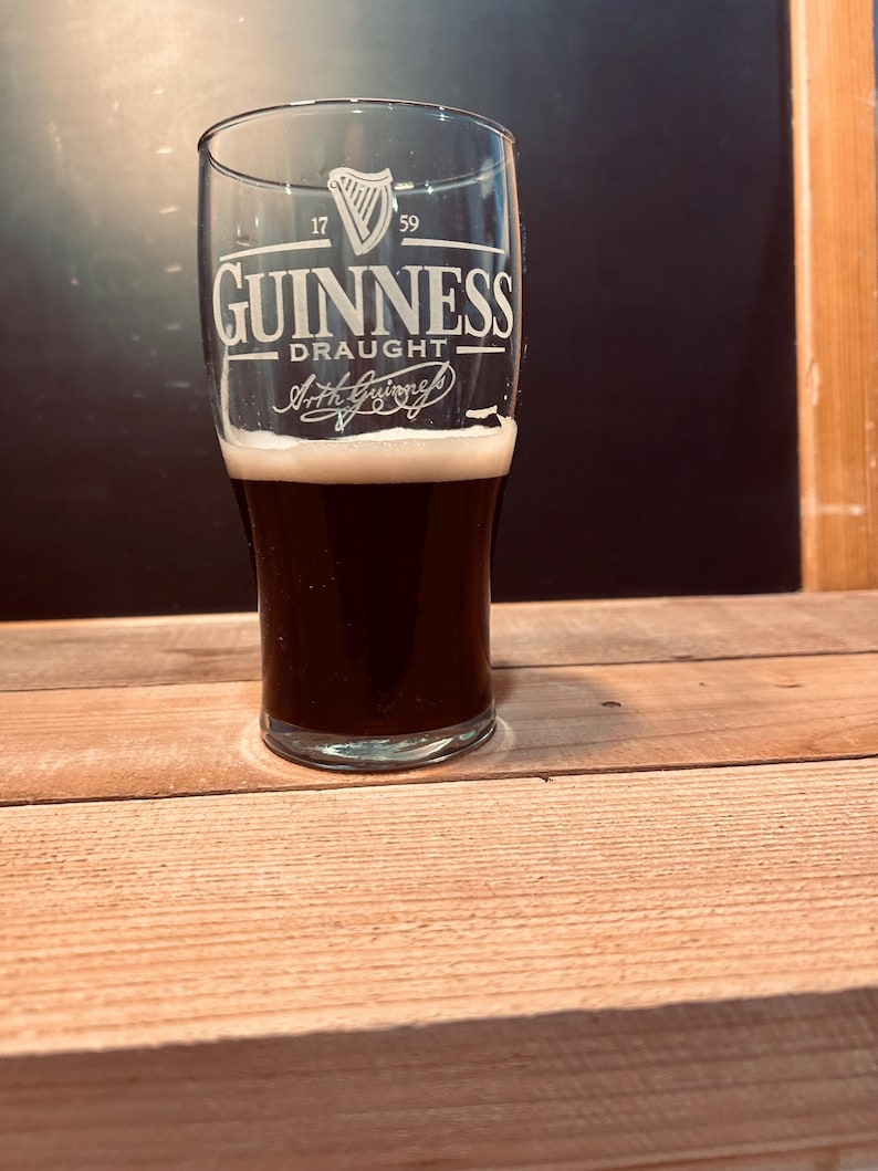 May include: A pint glass of dark stout beer with a creamy head, bearing the Guinness logo. The glass displays the harp symbol and the words "Guinness Draught". The glass is on a wooden surface.