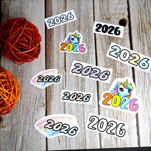 Könnte beinhalten: Eine Sammlung von Aufklebern und dekorativen Elementen mit dem Jahr "2026". Designs beinhalten die Zahl "2026" in verschiedenen Schriftarten und Farben, zusammen mit einer Einhorn-Illustration. Die Aufkleber sind auf einer Holzoberfläche angeordnet.