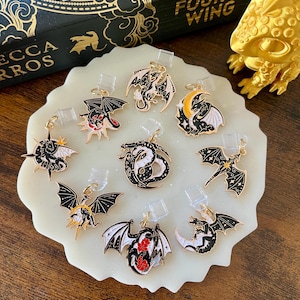 May include: A selection of dragon-themed charms in gold, black, white, and red. The charms are presented on a white, irregular-shaped surface. The charms feature diverse designs, including dragons with wings, moons, and stars. A book titled "Fourth Wing" is visible in the background.