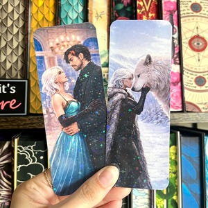 May include: Two fantasy-themed bookmarks. One features a couple in formal attire, the other a woman with a wolf. Both have a holographic sparkle effect. The background includes a shelf of decorative books.