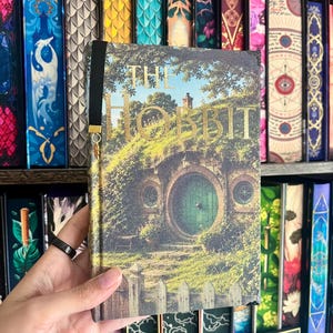 May include: A hardcover book titled The Hobbit featuring a printed illustration of a green round door set into a grassy hillside. The cover has a black ribbon bookmark with a small gold-colored charm attached to the end, resting against a bookshelf.