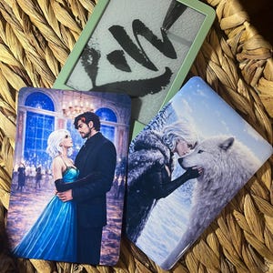 May include: Three rectangular cards with fantasy art and a green e-reader are displayed in a woven basket. The cards feature couples and a wolf. The e-reader displays black brushstrokes on a white background.