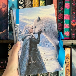 May include: A book cover depicting a woman and a white wolf in a snowy scene. The title "Fire Bound" is written in gold. A blue ribbon is tied around the book, which is held in a hand.