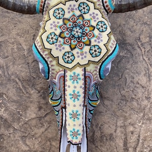 May include: A white cow skull decorated with colorful beads in a geometric pattern. The skull is adorned with turquoise, blue, gold, and purple beads. The skull is mounted on a wall.