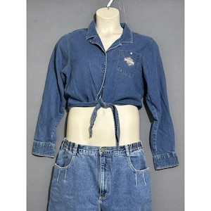 Może przedstawiać: Zestaw koszuli i szortów dżinsowych. Koszula z długim rękawem jest zawiązana w talii i posiada mały haftowany wzór na kieszeni. Szorty mają kieszenie i elastyczną talię. Zestaw jest w średnim niebieskim odcieniu.