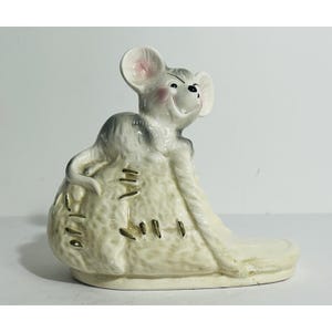 May include: A ceramic figurine of a smiling mouse with gray and white fur, perched on a beige sack. The mouse has pink ears and a black nose. The sack has stitched details and a flat base.