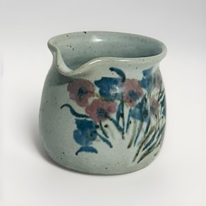 May include: A small, light blue ceramic pitcher with a spout. The pitcher is decorated with blue and pink floral designs. The handmade pottery has a speckled texture and a unique, organic shape, perfect for serving cream or sauces.