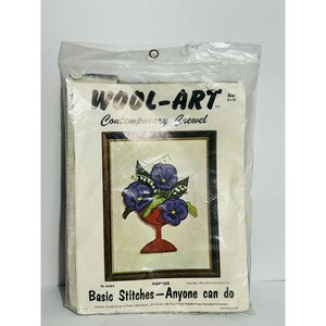 May include: A vintage "Wool-Art" embroidery kit featuring a framed floral design with purple pansies in a red vase. The kit includes the text "Basic Stitches - Anyone can do" and is packaged in a clear plastic bag.
