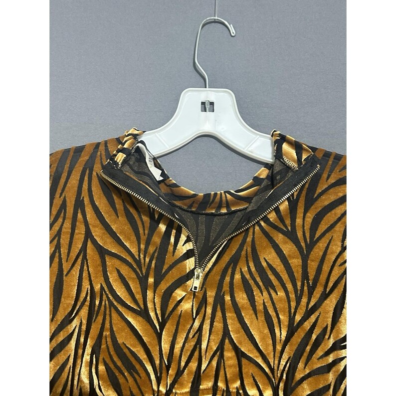 May include: A gold and black patterned top with a zip closure, hanging on a white hanger. The top features a repeating leaf-like design in black against a gold background. The zip is gold-coloured.