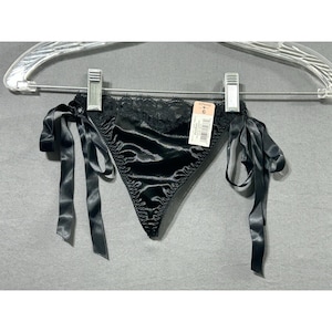 Pode incluir: Calcinha de cetim preta com acabamento em renda e laços de fita de cetim. A peça de roupa está pendurada em um cabide de plástico transparente sobre um fundo cinza. Uma etiqueta com um número está anexada.