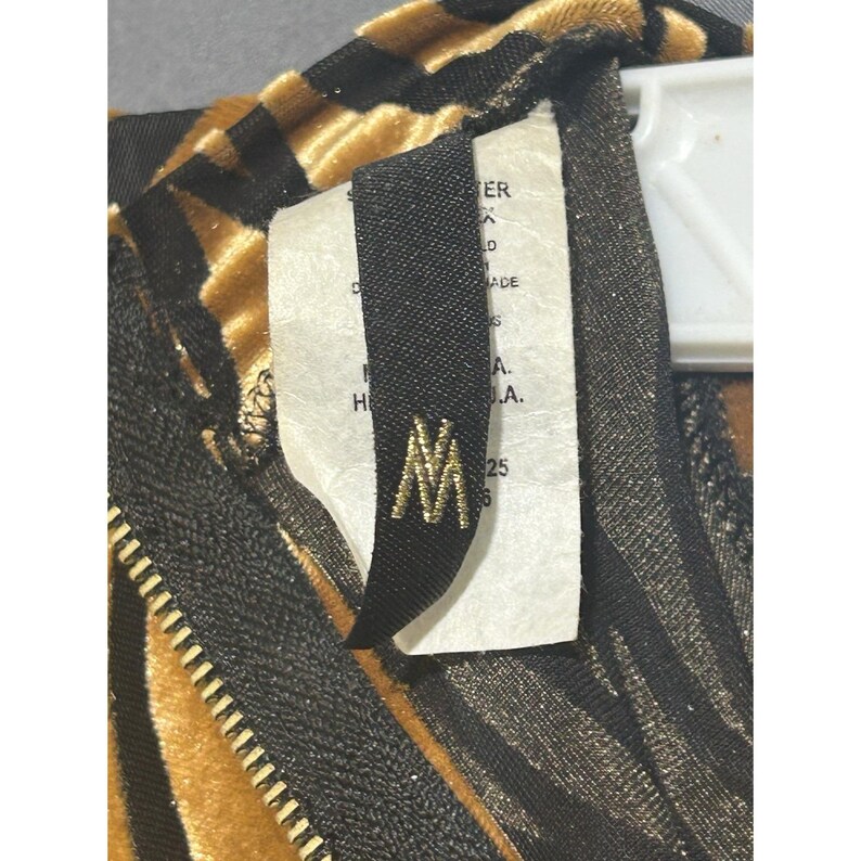 May include: Close-up of a garment with a gold and black striped pattern. A black tag with gold lettering and the letter "M" is attached. A zipper is visible on the left side.
