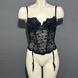 May include: Black lace corset with adjustable straps and garter straps. The corset features a floral pattern and underwire cups. The garment is displayed on a mannequin against a gray background.