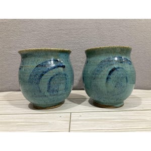 May include: Two handmade ceramic mugs. The mugs are a teal color with dark blue abstract designs. The rims are a lighter tan color. The mugs are sitting on a light wood surface.
