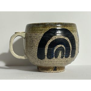 May include: A speckled ceramic mug with a rounded base and a handle. The mug features a dark blue, arched design on the side. The rim and handle are a lighter color, and the mug has a rustic, handmade appearance.