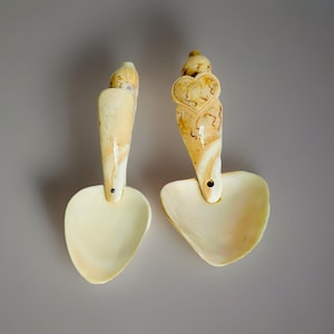 Op de afbeelding: Twee decoratieve lepels met hartvormige kommen en gesneden handvatten. De handvatten zijn crèmekleurig met bruine marmering, en een heeft een hartontwerp. De lepels staan tegen een grijze achtergrond.