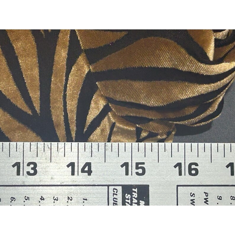 May include: Close-up of a fabric with a zebra stripe pattern. The fabric has gold and black stripes. A ruler is placed across the bottom of the fabric, with measurements in centimetres.