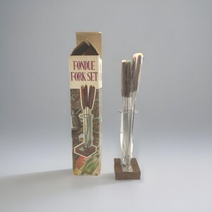 Puede incluir: Un juego de tenedores para fondue vintage en su caja original. La caja es de color beige con las palabras "FONDUE FORK SET" en rojo. El juego incluye varios tenedores con mangos de madera y un soporte de metal.