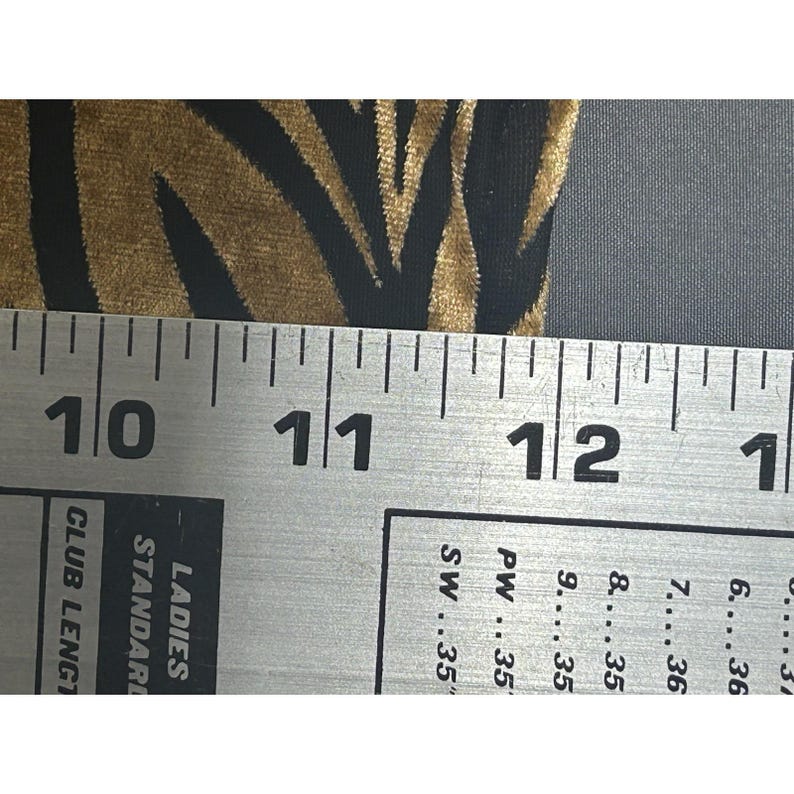 May include: Close-up of a fabric with a zebra stripe pattern in black and gold. A metal ruler is placed over the fabric, showing measurements from 25.4 cm to 30.5 cm. The ruler also displays text including "LADIES STANDARD" and "CLUB LENGT".