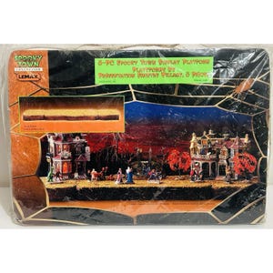Op de afbeelding: Een 3-delige Spooky Town displayplatformset van Lemax in de originele plastic verpakking. De doos heeft een zwarte achtergrond met een goudkleurig spinnenwebontwerp en toont een miniatuurdorp met twee huizen en diverse figuren op een basis.