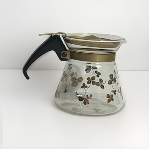 Peut inclure: Une cafetière en verre vintage avec une poignée noire et des accents dorés. Le verre transparent est décoré d'un motif de trèfles dorés. La cafetière a un couvercle et un bec verseur.