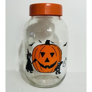 Könnte beinhalten: Ein klares Glas mit einem orangefarbenen Deckel, verziert mit einem Kürbis, einer Hexe, einem Skelett und Fledermäusen. Das Halloween-Glas eignet sich perfekt zum Aufbewahren von Süßigkeiten oder kleinen Gegenständen. Das Glas ist etwa 15 cm hoch.