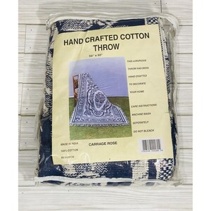 May include: A navy blue and white hand-crafted cotton throw, packaged in a clear plastic bag. The label reads "HAND CRAFTED COTTON THROW" and "50" x 60"" (127 x 152 cm), with care instructions and the "CARRAGE ROSE" design.