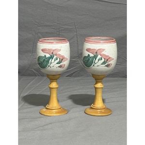 May include: Two decorative wine glasses. The glasses have a speckled white ceramic bowl with a pink rim and floral design. The stems are made of light-colored wood. The glasses are displayed on a gray surface.
