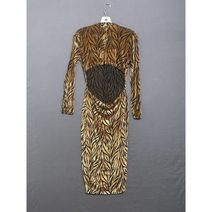 May include: A long-sleeved, figure-hugging dress with a leaf-like pattern in gold and black. The dress has a low back with a dark brown, textured panel. The dress is hanging on a white hanger against a grey background.