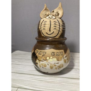 May include: A handmade ceramic cat-shaped jar, featuring a brown glaze and the words "FAT CAT". The cat has a smiling face, whiskers, and pointed ears. The jar is decorated with floral accents. A unique decorative item.