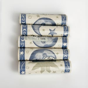 May include: Five rolls of wallpaper featuring a celestial pattern with dark blue crescent moons, stars, and swirls on a light beige background. Each roll has a dark blue border with white scrollwork and star motifs, and one roll includes a barcode label.