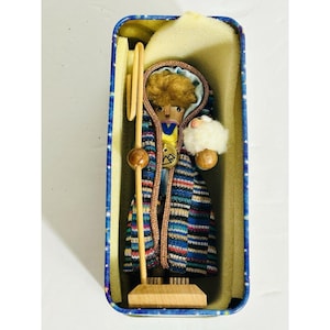 May include: A wooden shepherd figurine with a staff and a small sheep, inside a blue and yellow tin. The shepherd wears a colorful patterned coat and a hooded cloak. The figurine is approximately 4 inches tall.