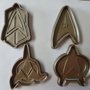 Könnte beinhalten: Vier hellbraune Ausstecher mit Star Trek-Insignien. Die Ausstecher zeigen das Starfleet Command, Starfleet und andere ikonische Symbole aus der Science-Fiction-Reihe. Ideal für thematische Backwaren.