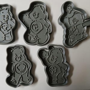 May include: Five gray plastic cookie cutters in the shape of Care Bears. Each cutter features a different bear design, including heart, cloud, and clover symbols. The cutters are arranged on a white surface.