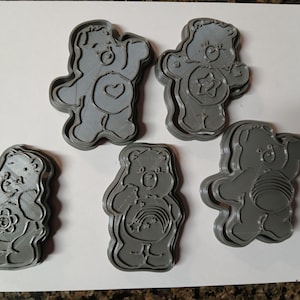 May include: Five gray Care Bear-shaped cookie cutters. Each cutter features a different Care Bear design, including a heart, star, flower, rainbow, and a ball of yarn. The cutters are made of a 3D-printed material and are set on a white surface.