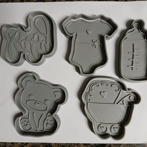 Op de afbeelding: Een set van vijf grijze plastic koekjesvormen met baby-thema, waaronder een woord in cursief schrift, een rompertje, een babyfles, een teddybeer en een kinderwagen. Elke vorm heeft een opstaande rand voor het snijden en interne details.