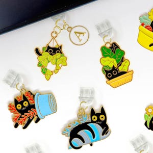 Può includere: Una collezione di ciondoli a tema gatto con vari disegni, tra cui gatti in vasi, piante e composizioni floreali. I ciondoli sono attaccati a clip trasparenti.